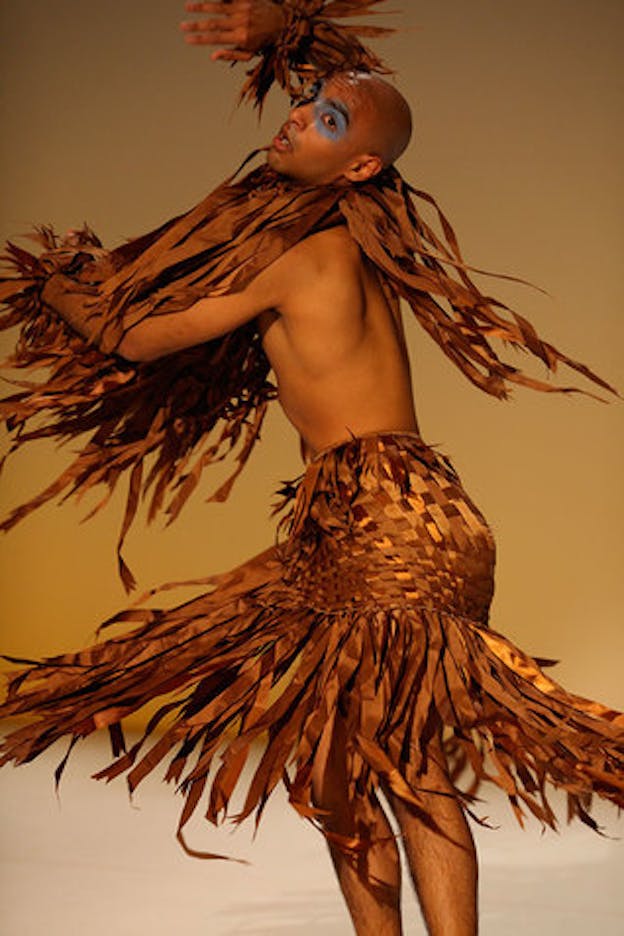 A performer in spinning motion wears an orange-brown midi skirt, a pair of wrist wraps, and a neckpiece all made of straps of lightweight fabrics weaved together. They hold one arm in the front and the other above the head while tilting their head to the front. The performer wears a baby blue eyeshadow that blends across their nosebridge. The stage has a beige floor and a sand color background.