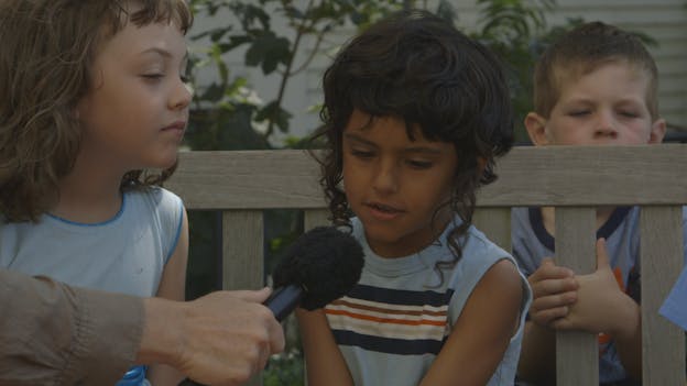 A young child wearing a light blue striped tank top is centered, sitting on a wooden bench outside with foliage in the background. They gaze downward as they speak into a black microphone held out to them by an adult on the left, whose body is out of frame. There is another child sitting to their right, who turns to listen as they speak. In the background is another child, holding onto the bench as they stand behind it. 