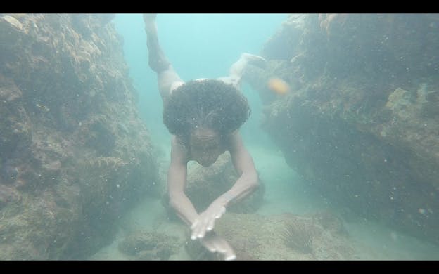 A performance still of nia love swimming towards the camera, nude, with her hands overlapped in front of her and her legs mid-kick behind her. Her eyes and mouth are closed tightly and she is nude. On either side of her (and below her) are big algae-covered rocks. The water is hued turquoise and the overall scene is slightly blurred. 