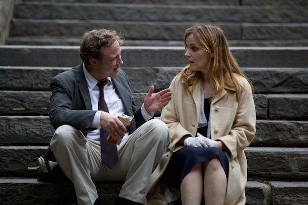 Two people dressed in gray and beige coats sit on stairs talking.