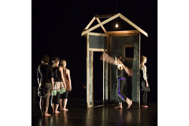 One performer stretches their arms up and down facing left in front of a simplified wooden house structure while another performer stands behind the structure facing back. A light bulb is hung from above to illuminate the interior of the house structure. In the front left, three performers are lined up according to their height facing towards the house structure behind them. All five performers are dressed in mismatched outfits. The performance is staged on a black floor against a black backdrop.