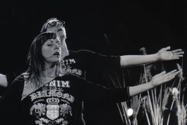 White and black photograph of two people standing behind one another with closed eyes and open arms, with the palms facing upwards.