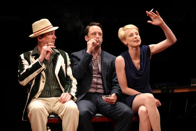 A performance still of three performers sitting on a cushioned black bench. On the far left, someone in a green and white striped suit jacket and a green striped shirt holds a cigar midway to their mouth and looks to the performer on the far right. In the middle, a performer in a black suit jacket and a purple shirt holds a cigar to their mouths and a glass ash tray on their knees. On the far right, a performer wearing a short blue dress raises their left hand as if about to ask a question. 