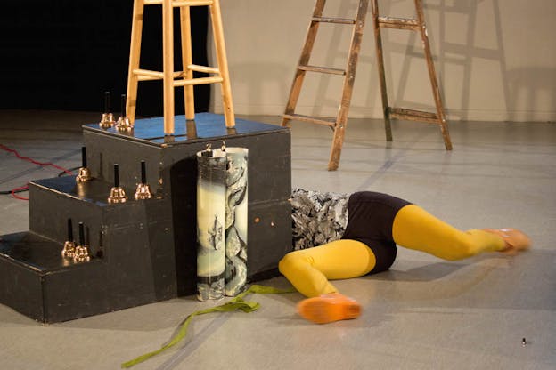 A performer dressed in black pants and yellow tights lays on the ground, the top half of their body covered by a black box stair with bells on the stairs and a chair on its top. 
