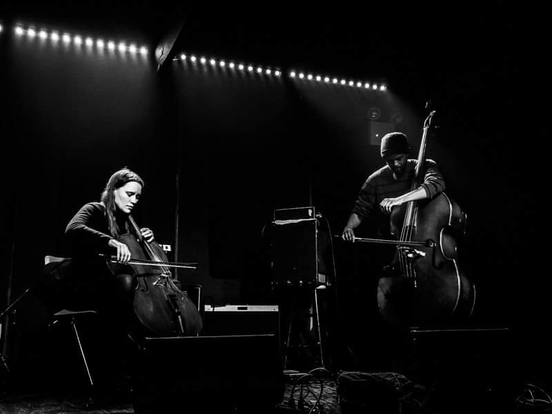 A black and white photo of Leila Bordreuil and Luke Stewart performing onstage. Leila is seated on the left side of the photograph playing cello and Luke is standing on the right side of the photograph playing upright bass with a bow.