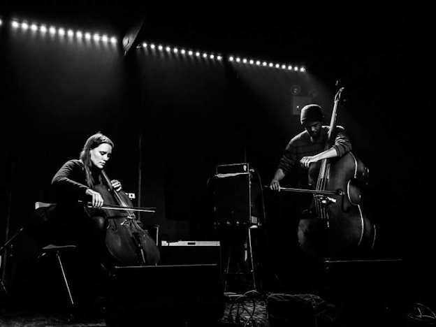 A black and white photo of Leila Bordreuil and Luke Stewart performing onstage. Leila is seated on the left side of the photograph playing cello and Luke is standing on the right side of the photograph playing upright bass with a bow.