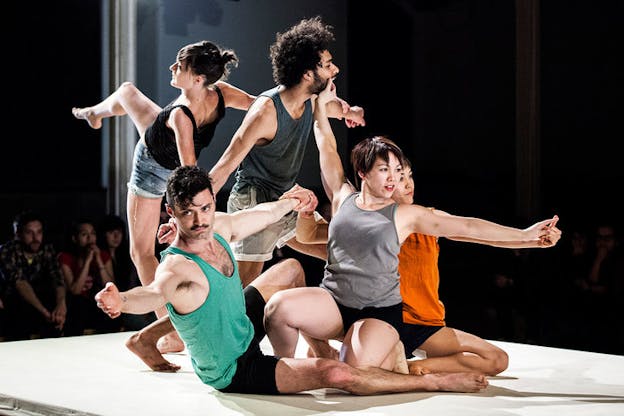 Performers in tank tops and shorts strike tangled poses atop an elevated stage: three positioned on the floor and two upright, they varyingly grasp each other by the jaw, the wrist, and the finger.