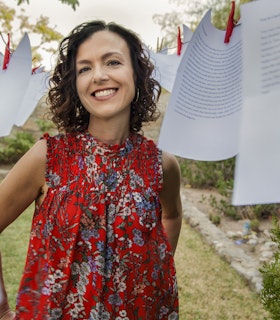 Rosa Alcalá is in the center of the image, smiling. Framing her face are pieces of paper fastened to a clothesline with a red clothespin. She wears a red patterned sleeveless top.