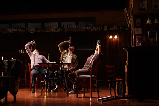 A performance still of three performers sitting at a table, leaning back and holding wine bottles to their mouths. They are in various street clothes. The room is dimly lit and there are other tables around them and a shelf filled with other wine bottles on the back wall. 