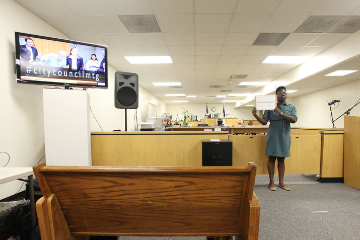 Empty meeting room except for one person in far back and one person holding a whiteboard with #citycouncilmtg written on it. To their left, a television portrays a group of suited people with the headline #citycouncilmtg. 