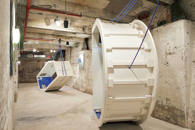 Two white structures open on two parallel sides hang from the ceiling by blue cords. Inside them blue bathroom tiles and a pair of slippers. 