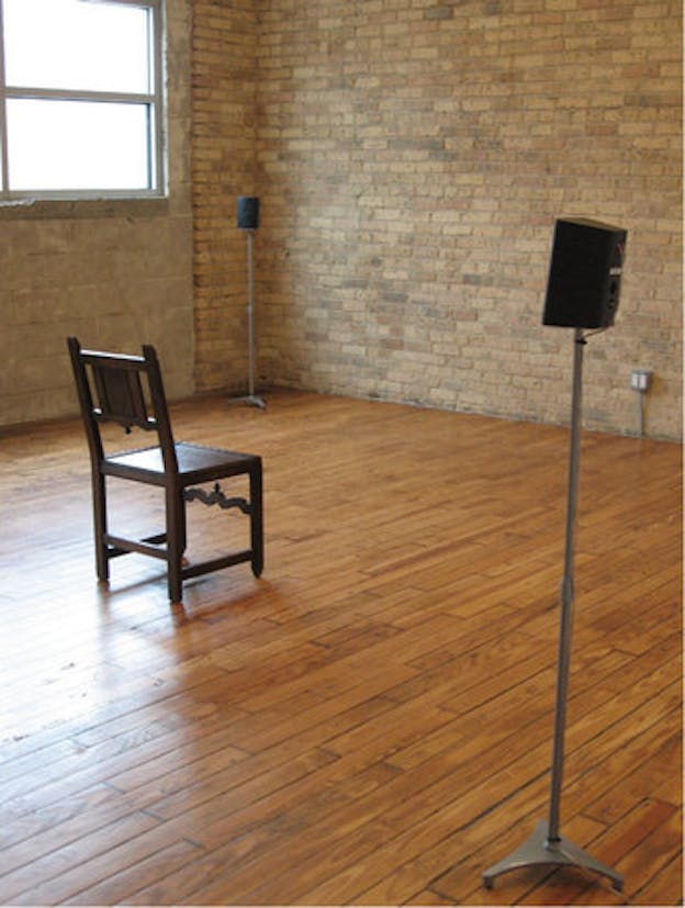 A single brown chair sits in the middle of the room facing a brown brick wall. Two speakers surround it. 