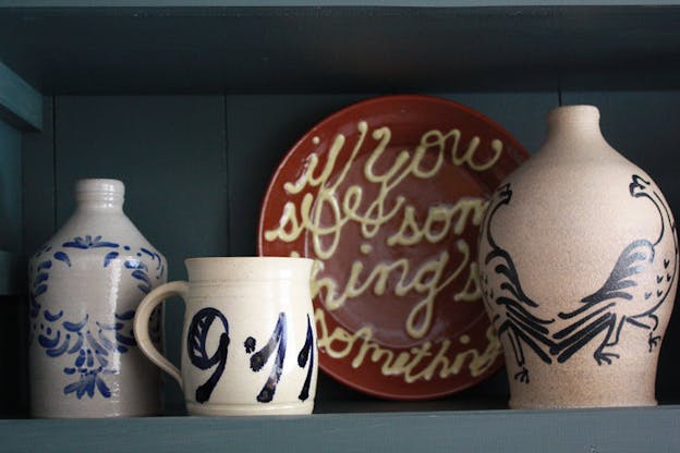 Four ceramic objects are arranged on a blue wooden shelf. On the far left, there is a grey jug with blue floral decoration. Next to that, there is a white mug with blue numbers 9 and 11. Behind that, there is a red plate with 