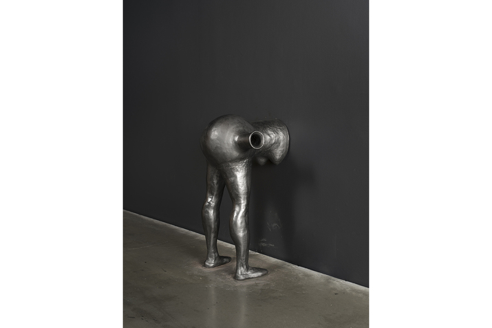 The silver statue of a figure crouching down on a wall with the features above the shoulders disappearing on the wall.