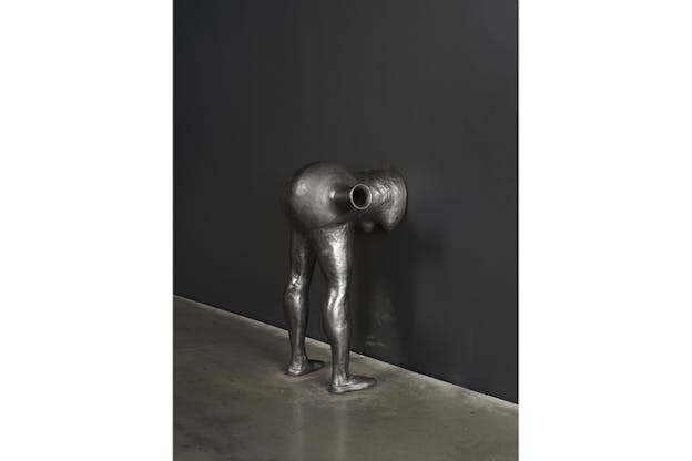 The silver statue of a figure crouching down on a wall with the features above the shoulders disappearing on the wall.