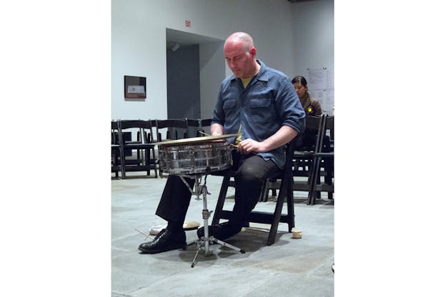 A person with their eyes closed sits in front of a single percussion instrument.