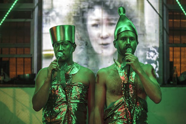 Tei Blow and Sean McElroy stand side by side, vocalizing into microphones and bathed in green light. They stand in sharp focus in front of a projection screen where there is a woman's face displayed out of focus. Both are wearing large golden headpieces and golden halter tops made of metallic, sparkly fabric. Blow's headpiece is flat-topped where McElroy's is tapered and cone-shaped with a rounded tip. 