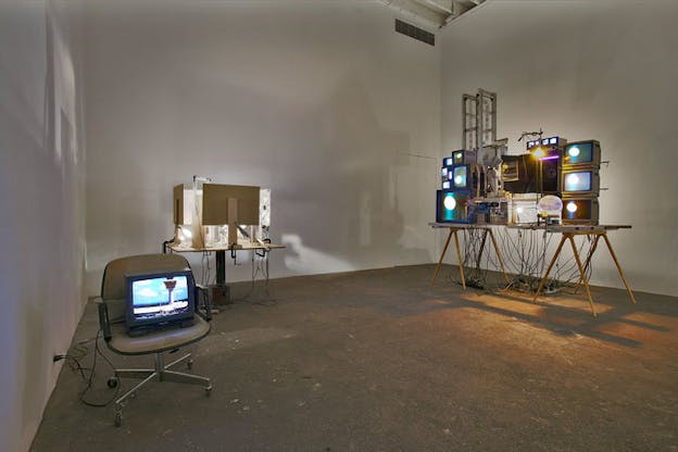 Several sculptures are situated within an otherwise bare white room. On the left, a television screen is on top of a the seat of a grey chair. This screen appears to show a monument in front of a blue cloudy sky. Behind this chair, there is a table with numerous computer desktops on top of it which are illuminated by white light. On the right, there is a table covered in stacks of differently sized television screens which project a blurred image of purple and blue light. 