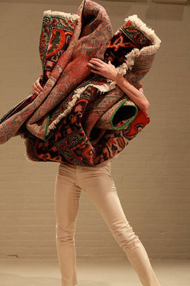 A performance still of Maria Hassabi in front of a cream colored brick wall. She wears pants that are the same cream color. In her arms, she holds a bundled carpet which covers her head and torso. 