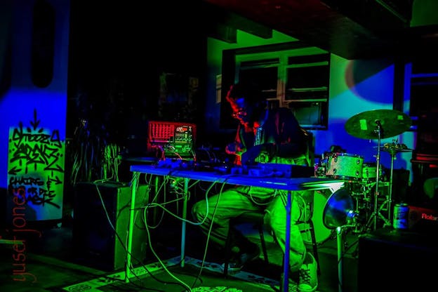 Luke Stewart sits in a dark room behind a mixing board, illuminated by multicolored green, red, and blue lights.
