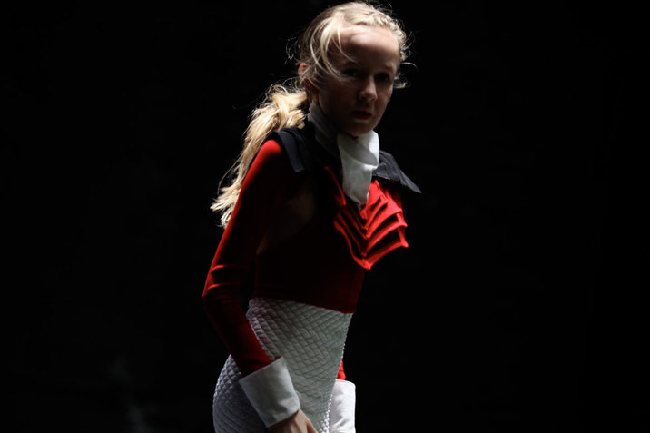A close up image of a performer dressed in a red shirt with ruffles at the neck and white shorts. They stand in the middle of a darkened space, looking intently at the camera.