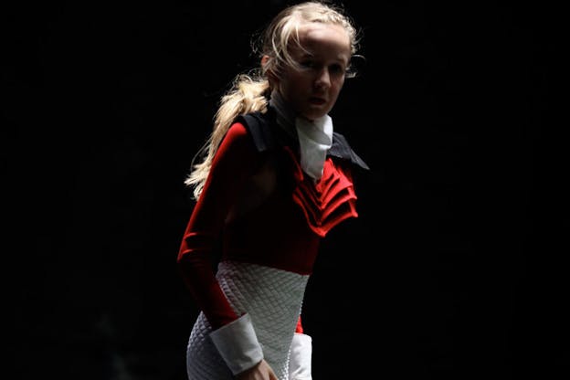 A close up image of a performer dressed in a red shirt with ruffles at the neck and white shorts. They stand in the middle of a darkened space, looking intently at the camera.