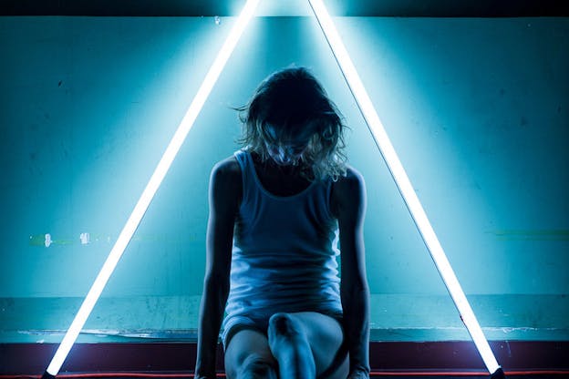 A performer sits in the center of a triangle made of blue neon lights. They clutch the surface beneath them and look down. They are wearing a white tank top. 