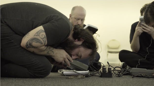 Nathan Young performs on the floor with three others in a well-lit, grey-carpeted room. Young is on the left half of the frame, kneeling with his head bowed towards the ground. He holds a wired electrical device to his face, his eyes are closed, wearing black jeans and a black t-shirt. The people behind him and to the right sit upright on the floor with closed eyes and crossed legs, also holding wired electrical devices. The black corded devices are plugged into a black power strip placed on the floor before Young.