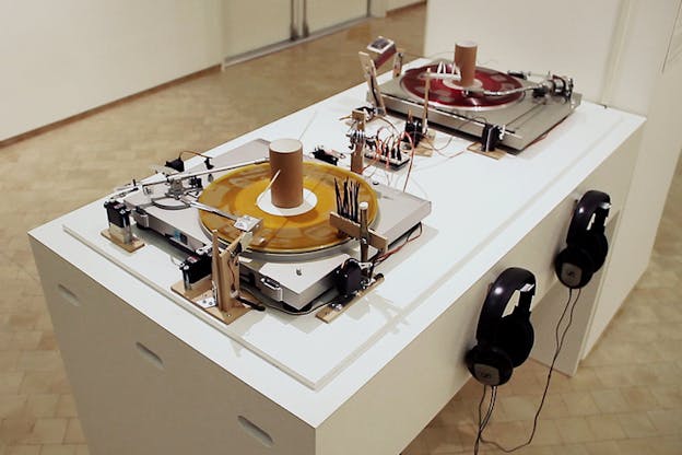 Two record players on a white table, each with a yellow and red record on them and various wooden equipment. On the side of the table under each record two headphones hang silver screwed hangers. 