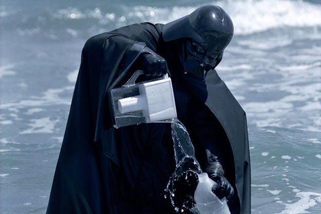 A person dressed as Darth Vader stands in ocean whitewash and pours water from a Brita into a plastic jug. 