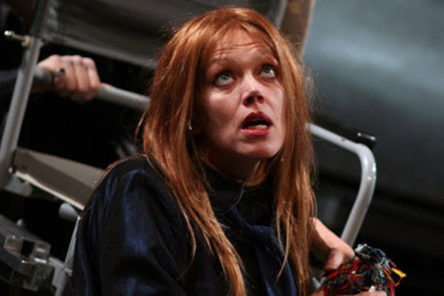 A close-up performance image of Kate Valk in front of a grey chair. She has long red hair and wears a dark blue top. In her hands, she holds a contraption made of red and silver wires. She looks up and opens her mouth as if mid-speech with an expression of exasperation on her face. 