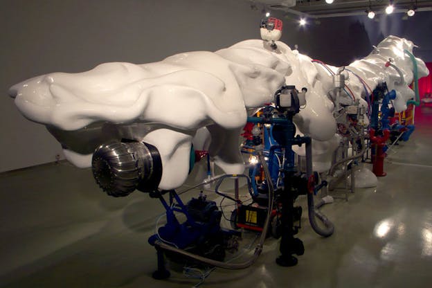 An installation image of a large mechanical object with various pipes and wires that are colored red, blue, and grey. A globular, organically curved white object rests on top of this. 