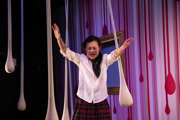 A person dressed in a white button down and red checkered skirt has their arms extended forward while they speak with closed eyes. Behind them white thin sacks hang in front of a pink and red wall.