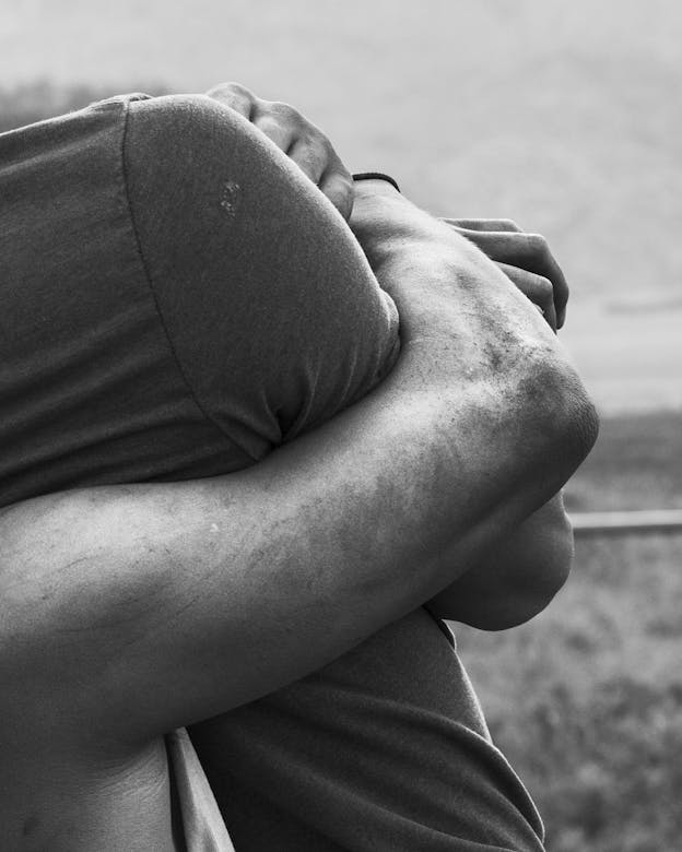 A black and white photo of a person's arm wrapped around another person, lifting them, in front of a grey, out of focus background. The lifting arm is soiled with dirt and the individual is wearing a tank top, only their arm and shoulder are visible. The person they are holding is wearing a t-shirt, their arm flush against their side, only their right shoulder and torso are visible. 
