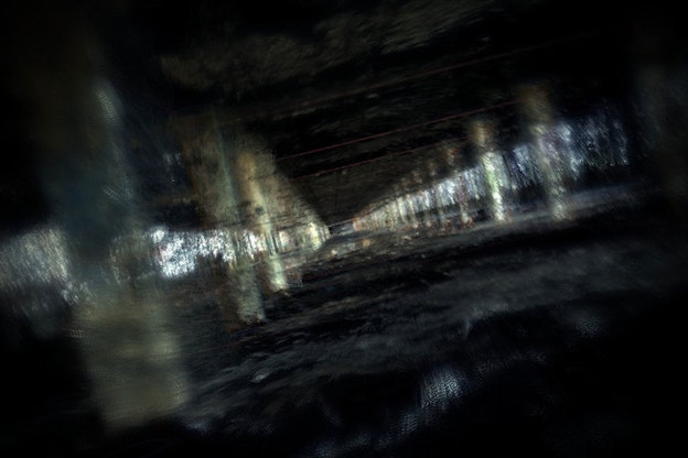 A blurry, tilted image of a dark room with many columns in two rows on either side. Behind the columns, there are glimpses of light blue. 