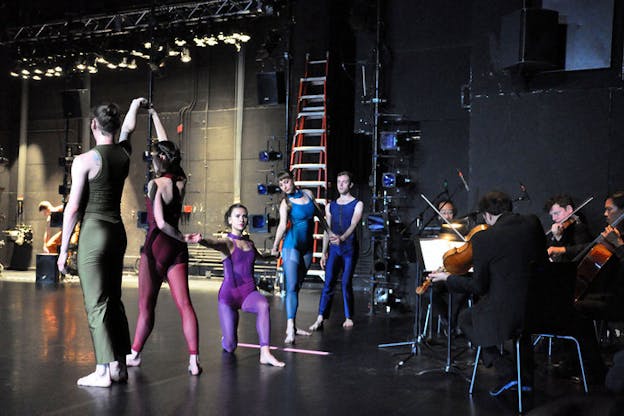 Five performers wearing monochromatic leotards and tights in varying hues of red, purple, and blue link hands and strike various poses onstage beside a quartet orchestra.