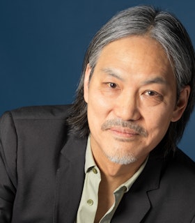 James Lo sits in sharp focus before a deep blue backdrop leaning towards the right. He smiles directly at the camera and is wearing a pale green button up shirt under a dark gray blazer.