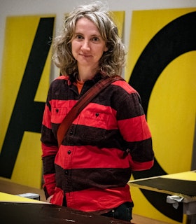 Sharon Hayes stands in the middleground, centered in a studio space with large yellow panels with black lettering in the background and on the table in front of her. She smiles directly at the camera, wearing a black and red striped button up shirt, the maroon strap of a crossbody bag over her shoulder. 