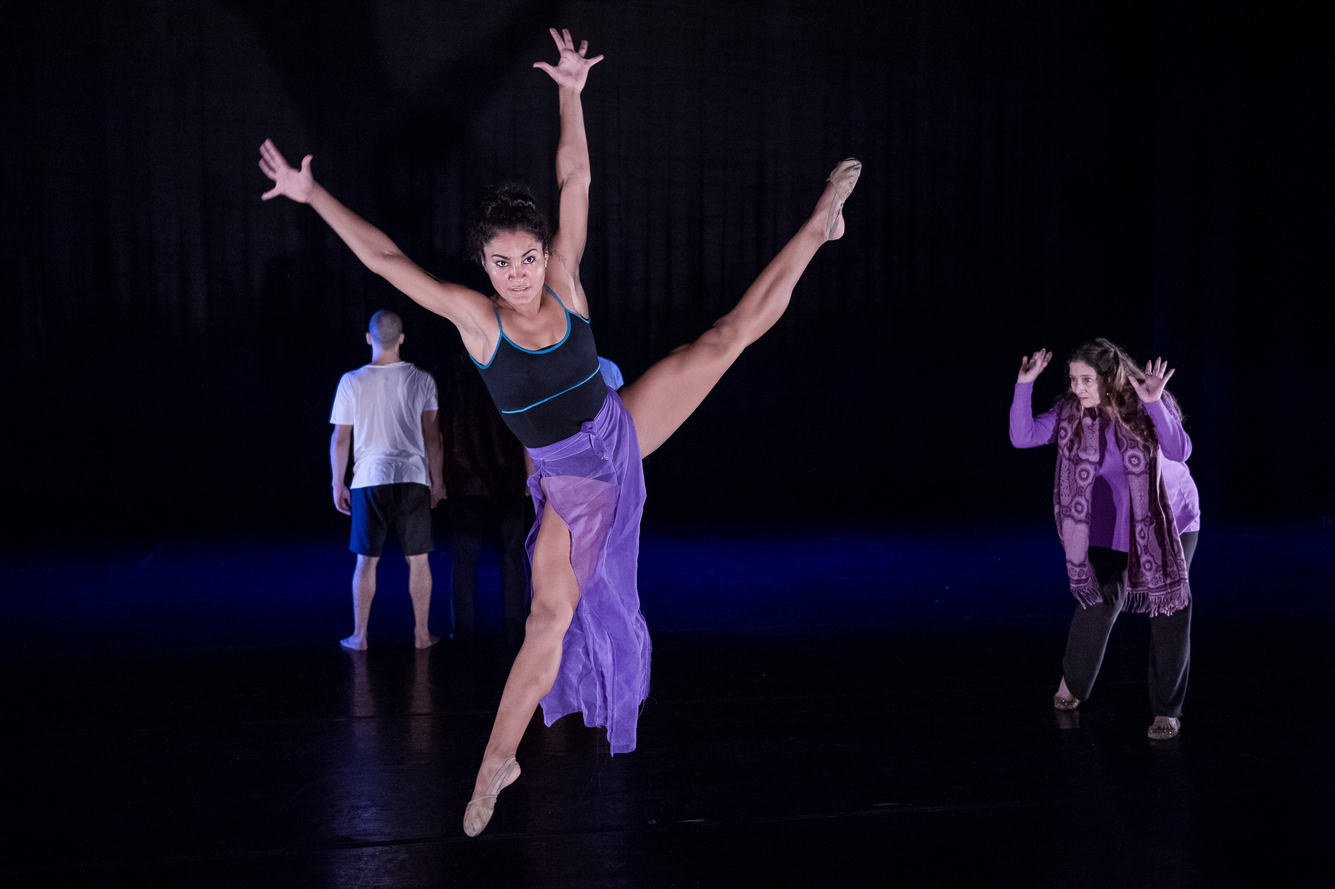 Three performers dance on a dark stage, illuminated from the front with one centered and leaping in the foreground, and the other two positioned in the background. Nayaret Ennovi is in the front and jumps in a wide split with her legs straight and arms raised in a Y, wearing a long purple skirt over a leotard. in the backround to the left, José Reyero stands, facing the back of the stage. On the right, Petra Bravo turns to look at Ennovi, hunched forward with her arms raised and hands framing her face, wearing a purple top, pants, and scarf.
