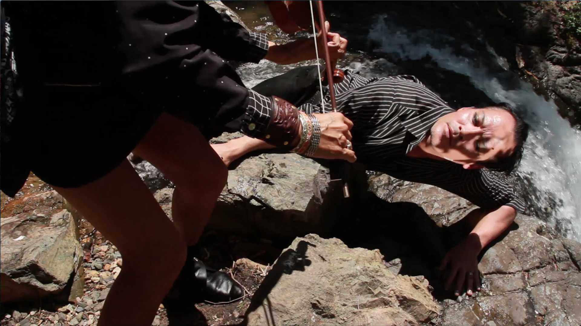 A performer lays down against stone. Besides them is a thin, rushing river. They look up towards Laura Ortman standing above them, leaning down towards them and playing the violin. The performer on the ground wears a black and whitestriped button up and eyeliner. Ortman wears a short skirt, black and white polka dot top, numerous bracelets, and black shoes. 