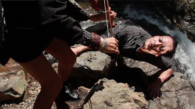 A performer lays down against stone. Besides them is a thin, rushing river. They look up towards Laura Ortman standing above them, leaning down towards them and playing the violin. The performer on the ground wears a black and whitestriped button up and eyeliner. Ortman wears a short skirt, black and white polka dot top, numerous bracelets, and black shoes. 