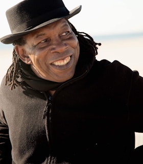 Newman Taylor Baker looks to his left, smiling widely and wearing a black zip up coat, a black scarf, and a black porkpie hat.