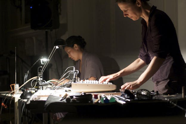 Two figures profile to the viewer work on a desk with scattered cables and other machineries in front of them. Two desk lamps are situated in front of each.