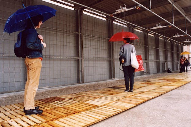 Four figures clad in dark clothes stand underneath transparent colorful umbrellas on a wooden runway. Rain falls atop them from machineries hanging from the ceiling.  