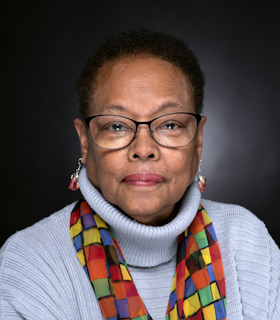 Cynthia Hawkins looks intently and directly into the camera. She wears glasses, rose-colored pendular earrings, a light blue turtleneck sweater, and a scarf with bright, multicolored squares.