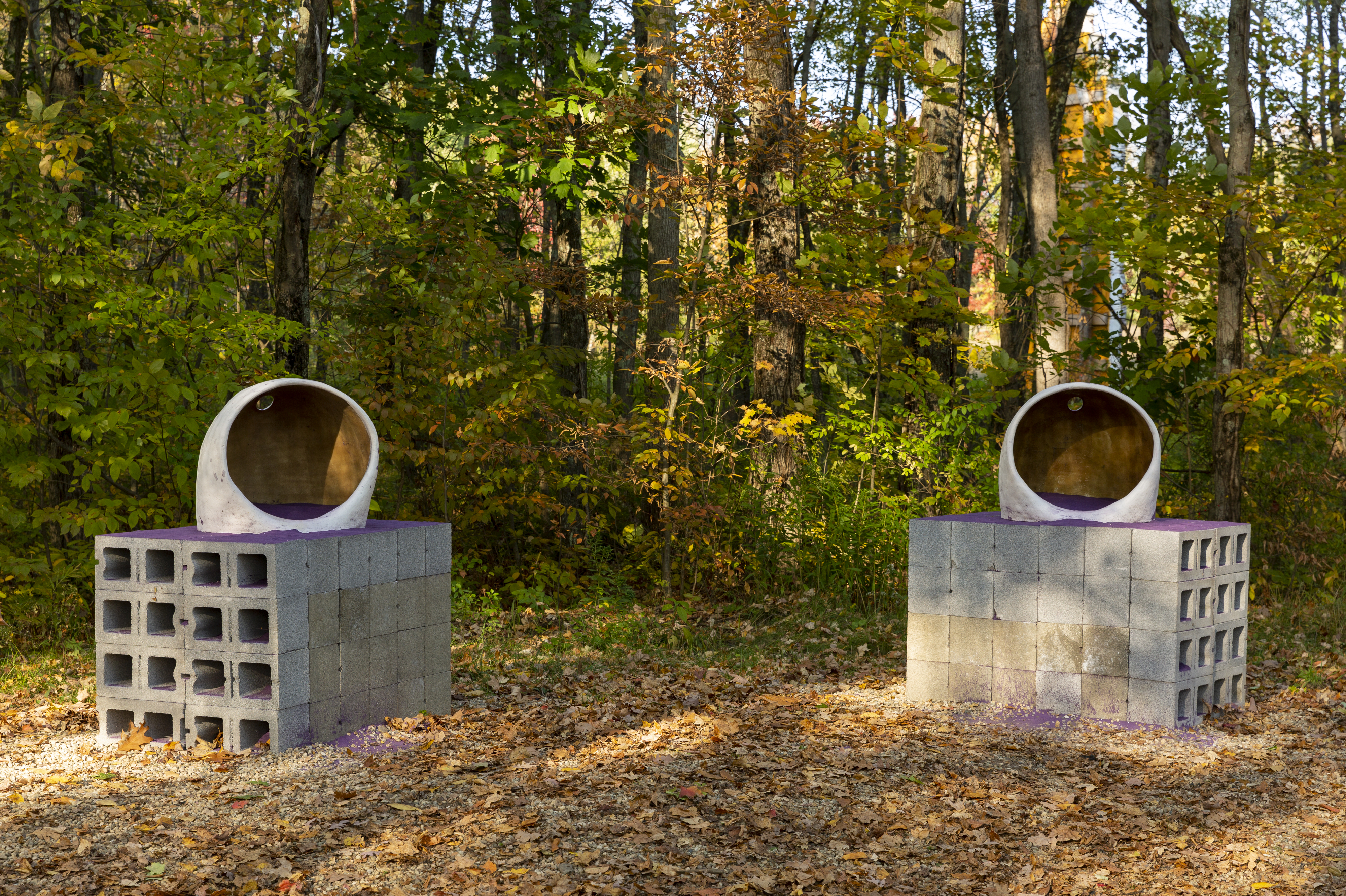Two sculptures sit beside each other in an outdoor wooded area. Both sculptures feature purple sand, a cinderblock pedestal, and an organic round shape.