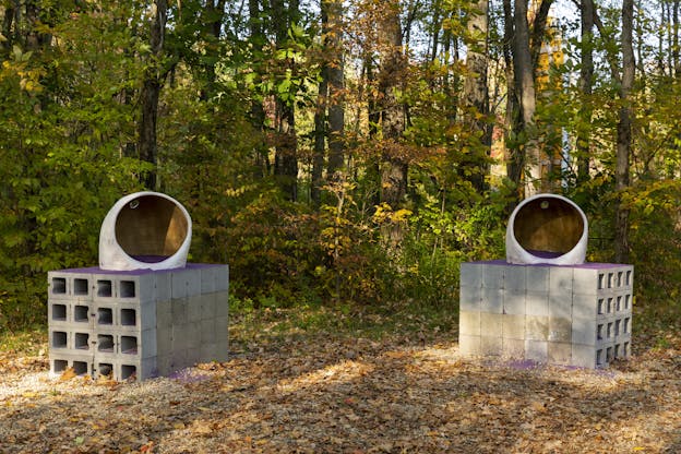 Two sculptures sit beside each other in an outdoor wooded area. Both sculptures feature purple sand, a cinderblock pedestal, and an organic round shape.