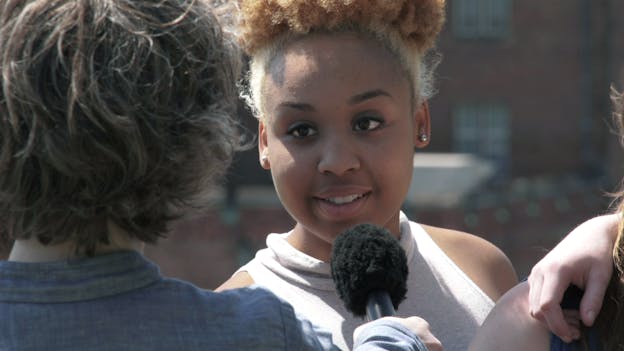 A closeup shot centering a young person with bleached hair as they speak into a microphone being in front of them. Sharon Hayes stands with her back to the camera on the left side of the frame wearing a grey collared shirt. In the right side of the frame, another person's shoulder is visible with an arm draped over it. 