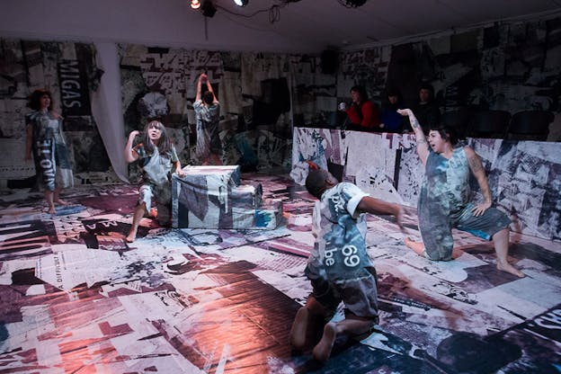 Performers dressed in clothing with newspaper pattern stand in a room covered by newspapers. Soft orange light illuminates on them as an audience watches them from behind a newspaper covered wall. 