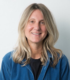 Portrait of Eve Fowler smiling and looking towards the camera. She wears a medium blue cardigan and a black shirt. She has medium length blonde hair and stands in front of a white background. 